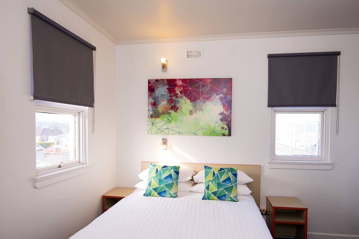 Une chambre à coucher comprenant un lit, deux tables de chevet, un tableau abstrait accroché au mur et deux fenêtres équipées de stores.