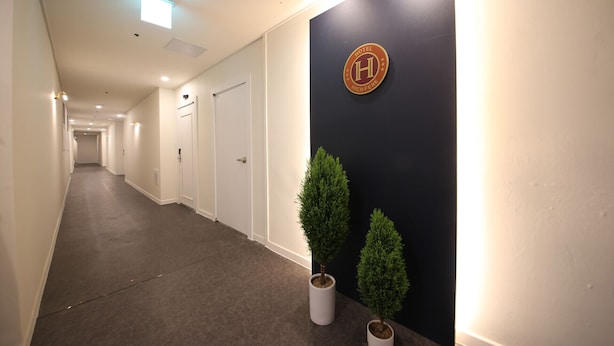 En hotellkorridor med en mörk vägg, en skylt med en logotyp i form av bokstaven ”H” och två krukväxter.