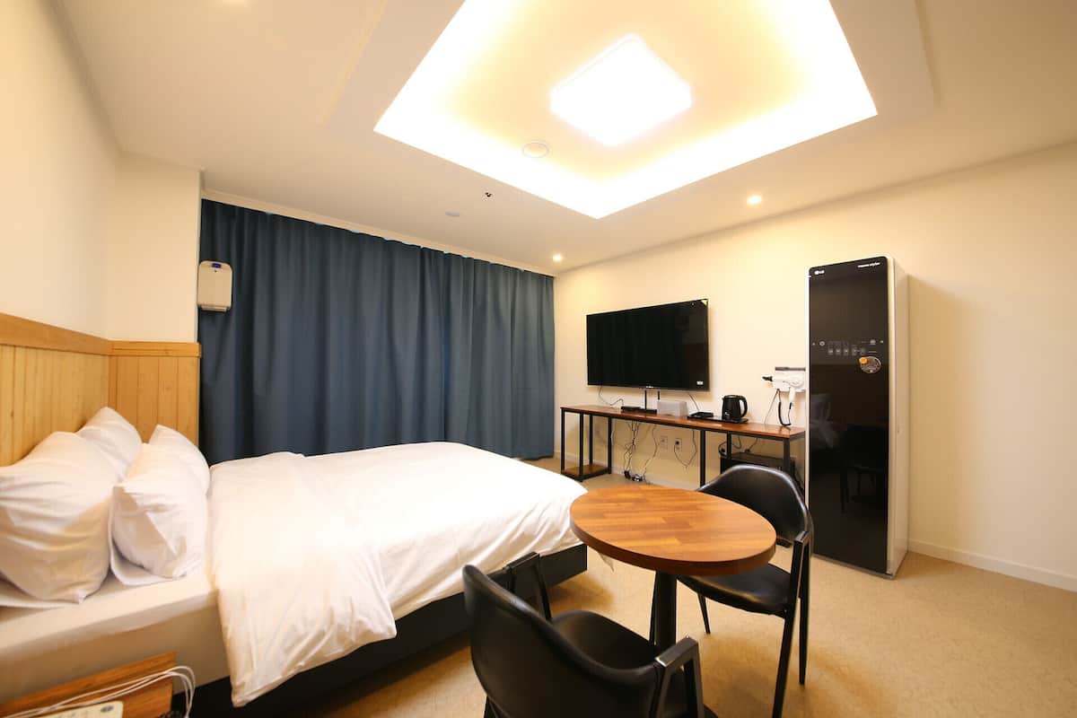 A hotel room with a bed, a desk with a flat-screen TV, a refrigerator, and a ceiling light fixture.
