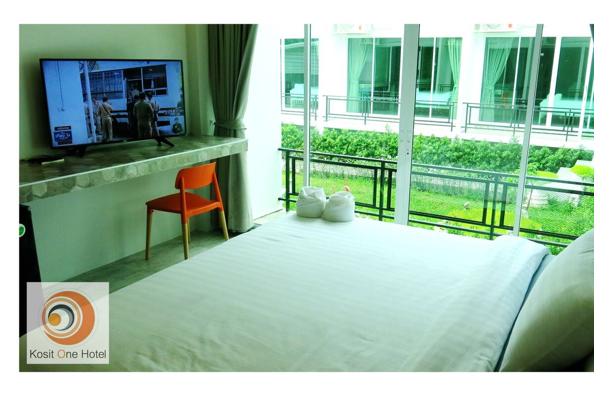 A hotel room with a bed, a TV on the wall, a chair, and a balcony with a view of greenery.