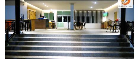 Un hall de hotel con mostrador de recepción, una escalera que sube al piso superior y una zona de estar con sillas y mesas.