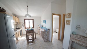 A kitchen with a refrigerator, wooden cabinets, a dining table with chairs, a hanging light fixture, and a view into another room.