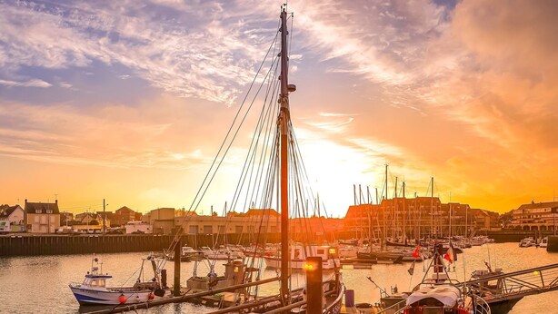 A harbor with boats docked, a bridge, and buildings in the background during sunset.