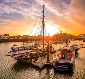 Un port avec des bateaux amarrés, un pont et des bâtiments en arrière-plan, le tout baigné par les couleurs du coucher de soleil.