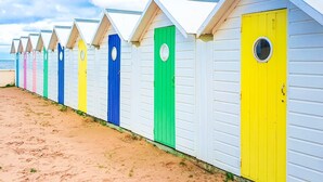 A row of beach huts with white exteriors and colorful doors, lined up along a sandy beach.