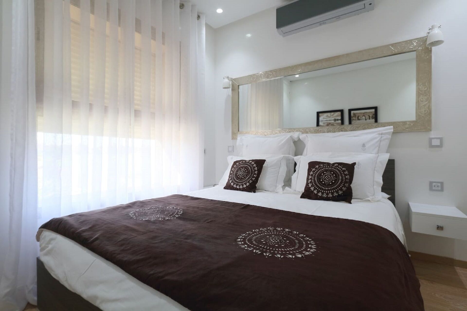 A bedroom with a large bed, white bedding, a decorative mirror, and a window with sheer curtains.