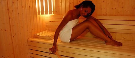 A person sitting on a wooden bench in a sauna room with a wooden bench and a window with blinds.