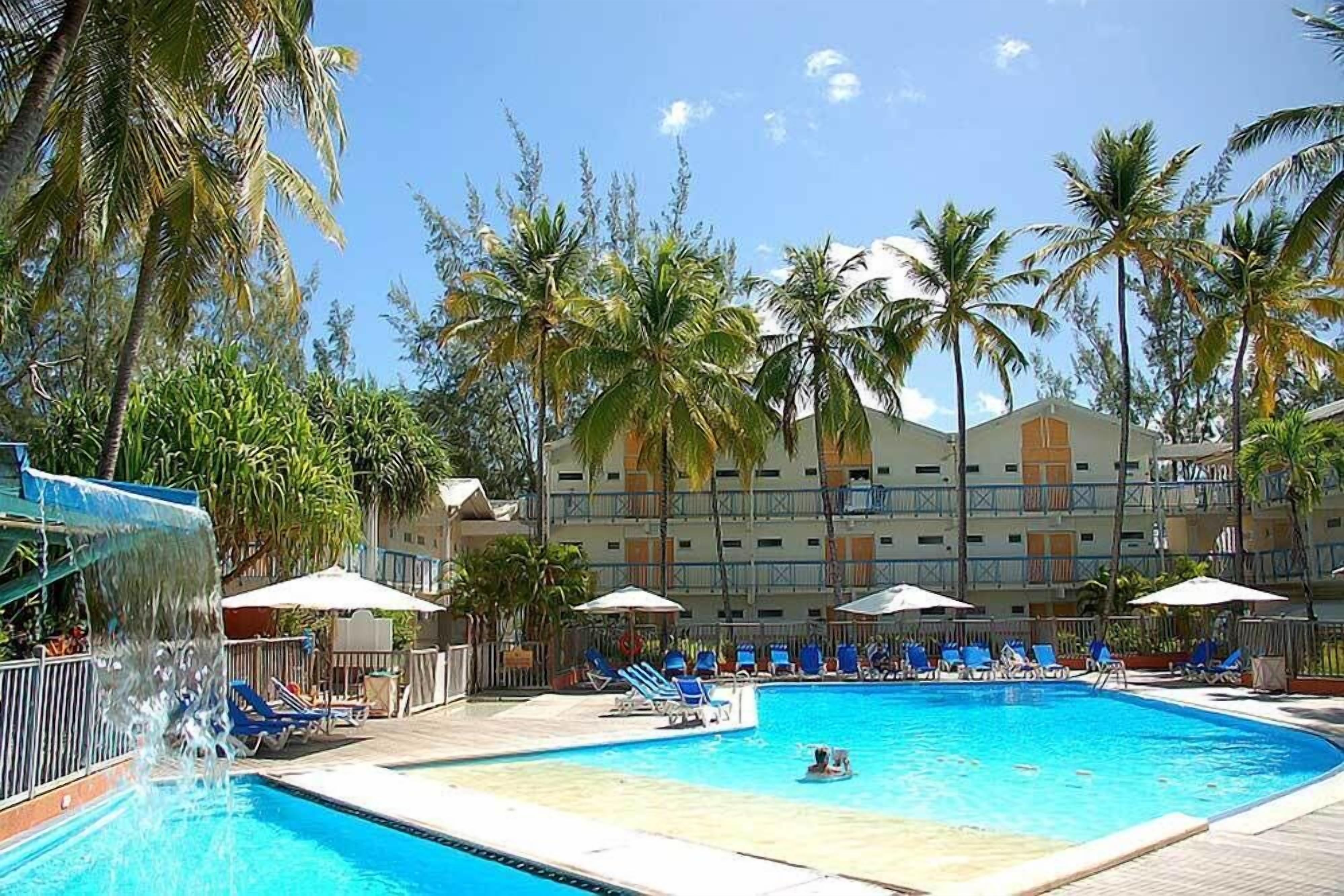 A swimming pool with lounge chairs and umbrellas, surrounded by palm trees and a hotel building in the background.