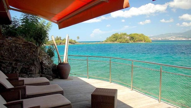 Een terras met rieten meubilair en uitzicht op turquoise water en een eiland in de verte.