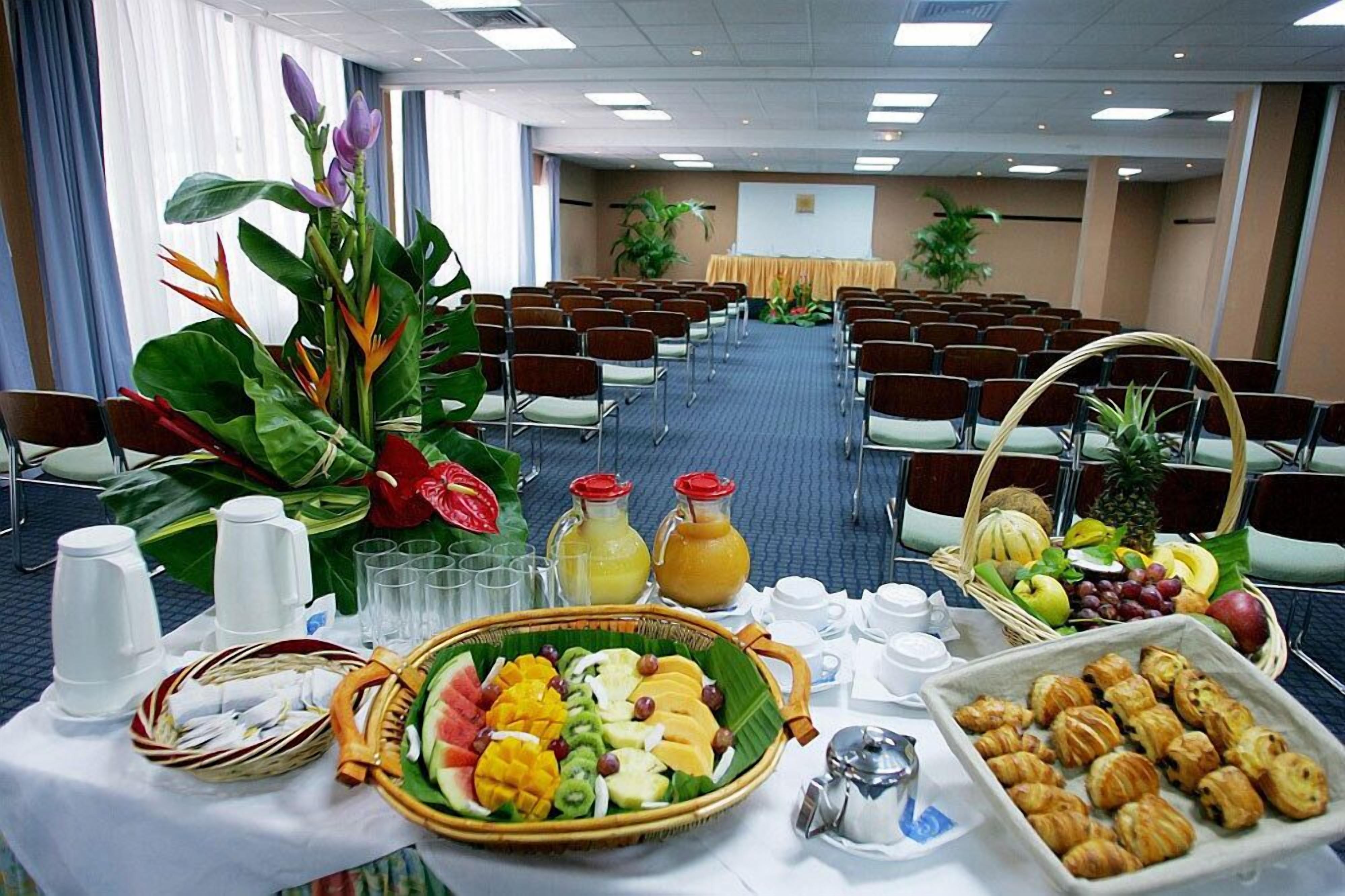 Ein Konferenzraum mit Reihen von Stühlen, einem Buffet mit Früchten, Gebäck und Getränken sowie einer Projektionsleinwand im Hintergrund.