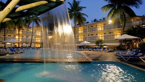 Zwembadgedeelte van een hotel met een waterval, ligbedden en palmbomen.