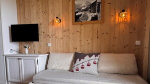 A cozy room with a wooden wall, a white sofa, a flat-screen TV, and a small table with a bowl of decorative items. - T3 for 6/7 people with direct access to the slopes, beautiful view, summer swimming pool, shops (Valmeinier)