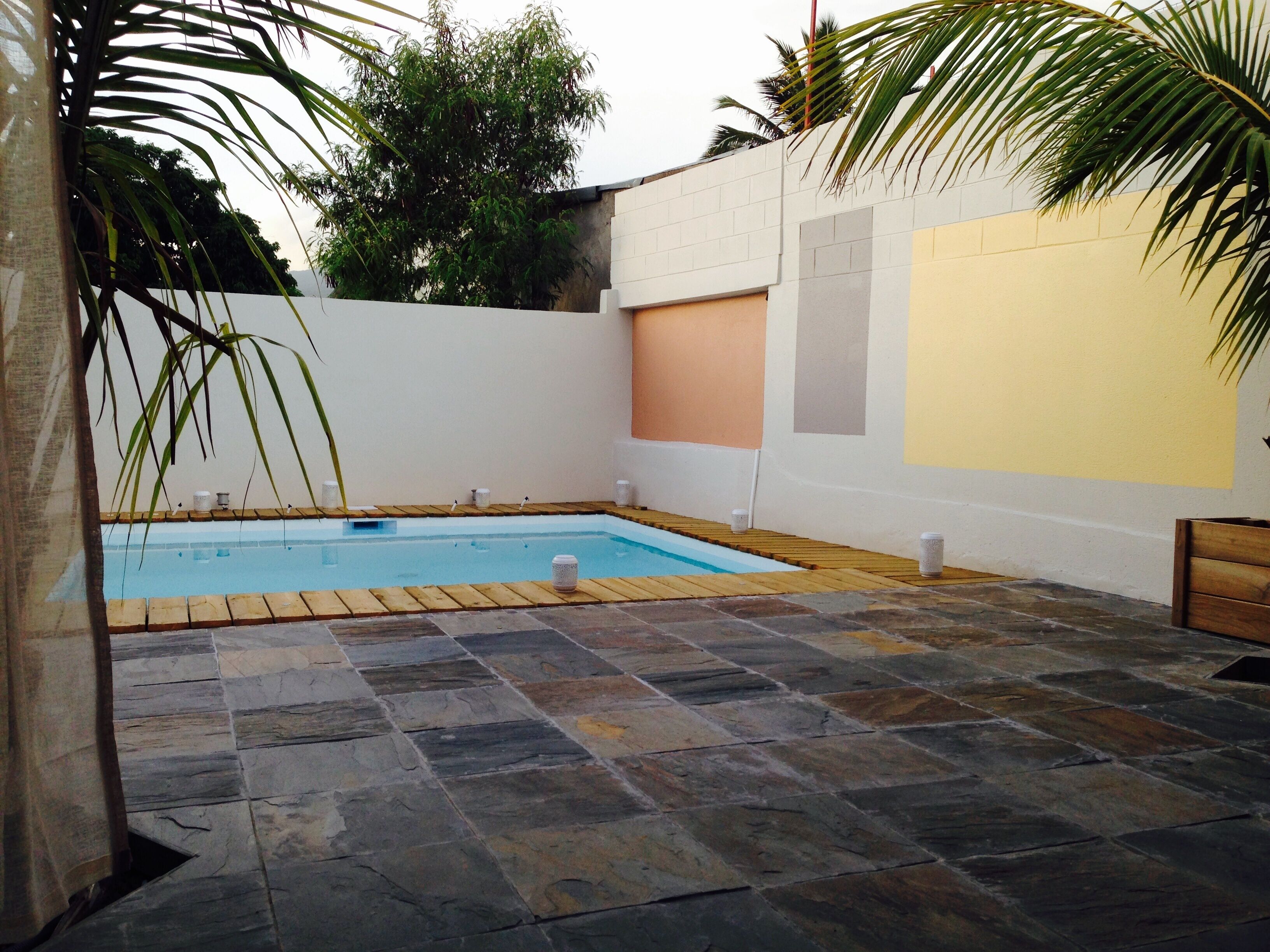 A swimming pool with stone tiles, surrounded by a white wall with a yellow section, and palm trees.