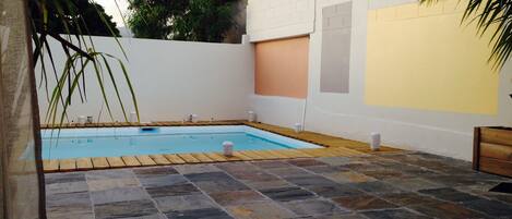 A swimming pool with stone tiles, surrounded by a white wall with a yellow section, and palm trees.