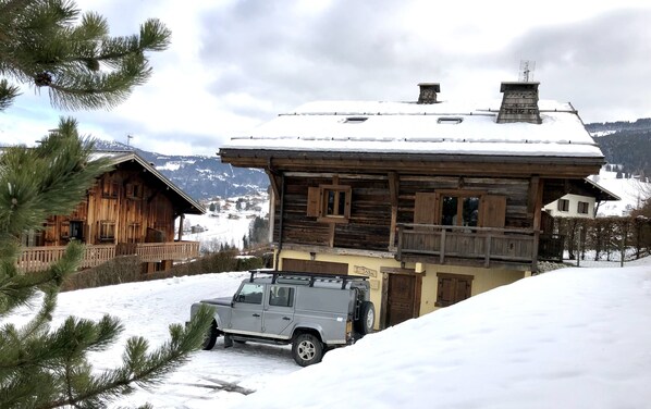 Une cabane en bois dotée d’un balcon, un véhicule garé et un paysage enneigé avec des montagnes en arrière-plan.