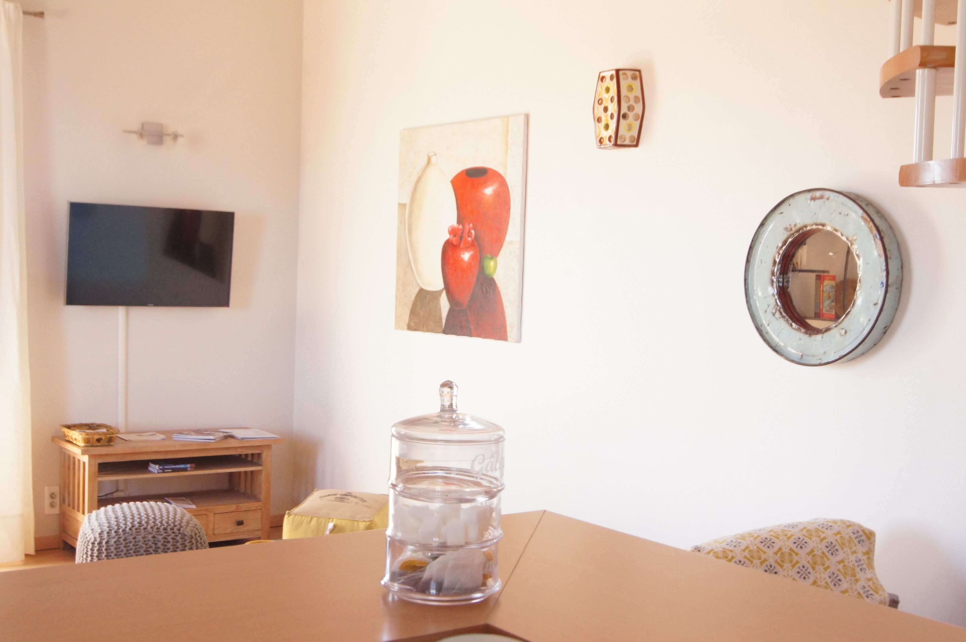 A room with a TV mounted on the wall, a wooden desk with a chair, a large jar on the table, and a painting.
