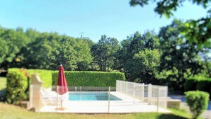 Une piscine entourée d’une clôture blanche, de verdure et d’un parasol rouge.