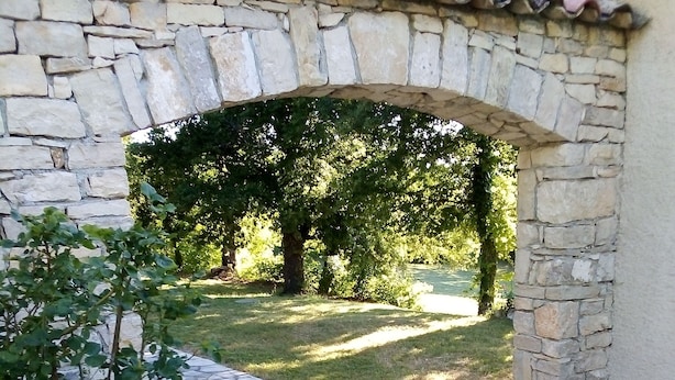 Un arc en pierre mène à un espace verdoyant où se trouvent une plante en pot et un petit pot posé sur une corniche en pierre.