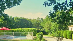 Un espace piscine agrémenté d’un parasol rouge, une piscine entourée d’une clôture et un petit jardin avec des arbustes soigneusement taillés.