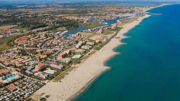 Vista aérea de un complejo turístico frente al mar con propiedades en la playa, un puerto deportivo y una playa con sombrillas.