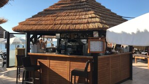 Un bar en la playa con techo de paja, mostrador de madera y dos sillas de mimbre.