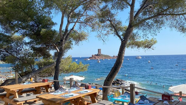 Una terraza de madera con mesas de picnic y bancos, con vista a una playa con barcos y una isla rocosa al fondo.
