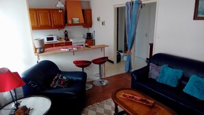 Una sala de estar con un sofá, dos taburetes rojos, una mesa de centro y una pequeña mesa con un plato. Hay una zona de cocina con armarios, un microondas y una cafetera. Una cortina azul cuelga en la entrada.