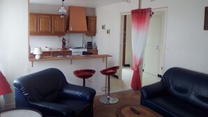 Una sala de estar con dos sofás de cuero azul, una mesa de centro y una cortina roja. Se ve una cocina con gabinetes de madera y una estufa al fondo.