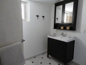 Une salle de bain avec un lavabo blanc, un meuble noir et un miroir au-dessus. Il y a deux lampes fixées au mur et une fenêtre avec une serviette accrochée à la porte.