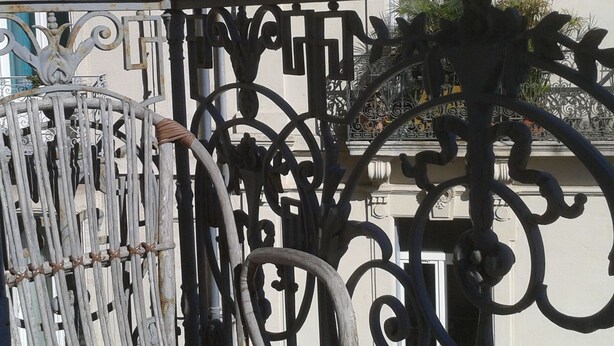 A wrought iron balcony gate with intricate designs, a white building with a window and satellite dishes in the background.