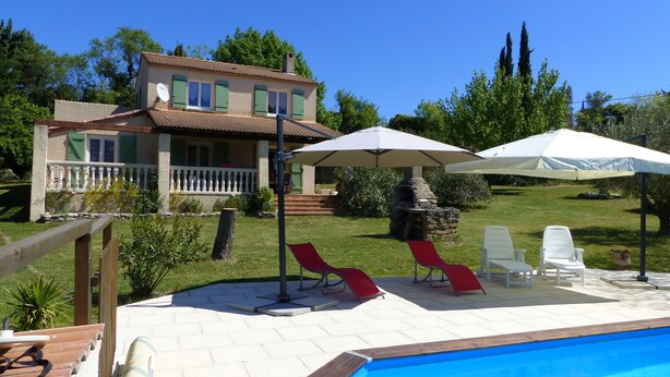 A swimming pool with lounge chairs and umbrellas, a stone fireplace, and a house with a balcony and green shutters.