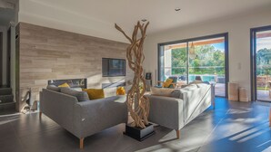 A modern living room with two grey sofas, a wooden sculpture, and a large window offering a view of greenery. - Incredible 5* villa near Cala Rossa (Porto-Vecchio)