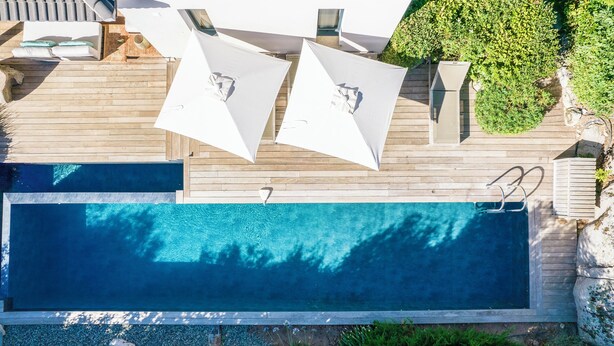 A rectangular swimming pool with a wooden deck, two white umbrellas, and a small garden area with plants and a rock feature. - Incredible 5* villa near Cala Rossa (Porto-Vecchio)