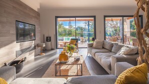 A modern living room with a large sofa, a coffee table, and a TV mounted on the wall. There is a sliding glass door leading to an outdoor patio area. - Incredible 5* villa near Cala Rossa (Porto-Vecchio)