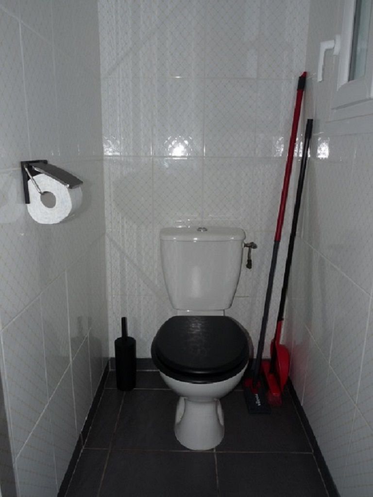 Een kleine betegelde badkamer met toilet, toiletrolhouder en een bezem die tegen de muur staat.