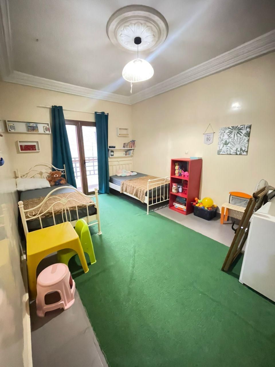 Una habitación con dos camas, una estantería roja, un taburete pequeño y un ventilador de techo.