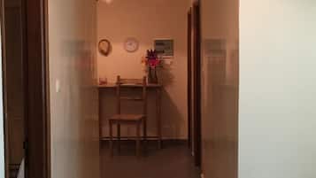Un pasillo estrecho con una puerta de madera a la izquierda, una pequeña mesa con una silla y un florero en el centro, y un par de zapatos en el suelo a la derecha.