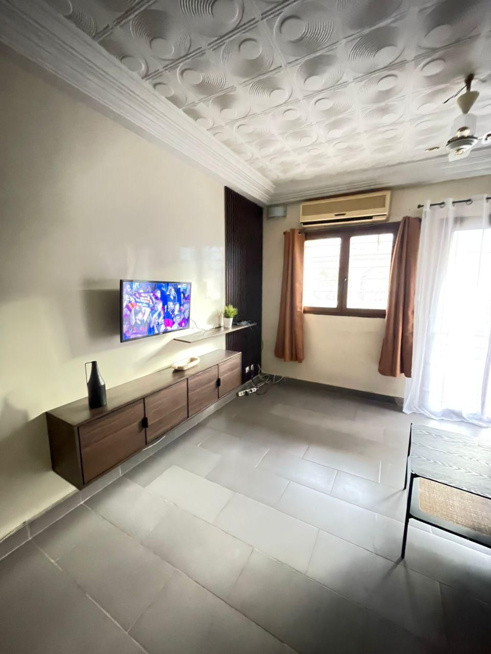 Una habitación con una televisión de pantalla plana montada en la pared, un aparador de madera, una ventana con cortinas y un techo con molduras decorativas.