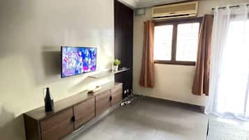 Una habitación con una televisión de pantalla plana montada en la pared, un aparador de madera, una ventana con cortinas y un techo con molduras decorativas.