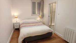 A narrow bedroom with a bed, two bedside tables with lamps, a window with curtains, and a mirror.