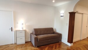 A room with a wooden floor, a brown sofa, a white bedside table, and a white wardrobe.