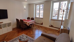 A living room with a flat-screen TV, a dining table with chairs, a sofa, and a large window with a view of buildings.