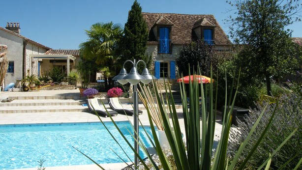 A pool area with a clear blue pool, a pergola with umbrellas, and a house with blue shutters and a tiled roof. - Superb gîte with private heated pool, air-conditioned rooms  (Pomport)