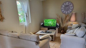 A living room with a beige sofa, a smaller armchair, a coffee table with a decorative plate, a television, a wall clock, and a lamp. - Superb gîte with private heated pool, air-conditioned rooms  (Pomport)