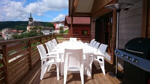 Una terraza de madera con una mesa, sillas, una parrilla y vistas a una iglesia y casas al fondo.