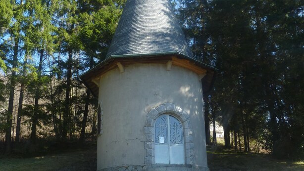 Una pequeña capilla de piedra con forma cónica y una cruz en la parte superior, rodeada de árboles.