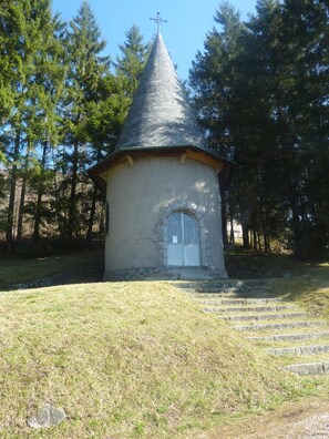 A small, conical stone chapel with a cross on top, surrounded by trees.