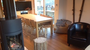 Ein gemütliches Wohnzimmer mit Kamin, einem Esstisch mit Stühlen, einem schwarzen Ledersessel und Blick auf einen Balkon mit Steinmauer.