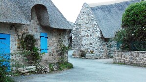 A narrow street with stone buildings, one with a blue shutter and another with a thatched roof.
