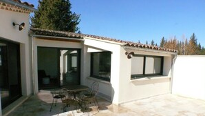 Une terrasse extérieure avec une table et des chaises, de grandes fenêtres et un sol carrelé.